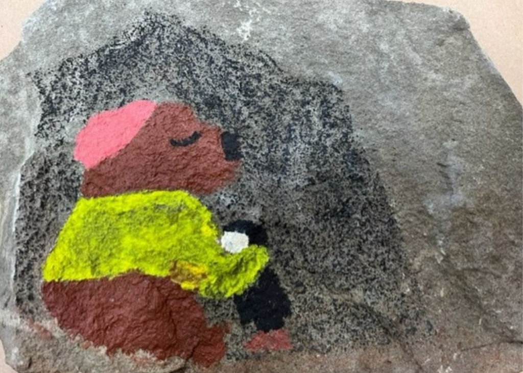This is a colorful painting on a gray stone. A brown bear wearing a yellow top and a pink hat is hugging a black penguin with a white face and brown feet 