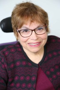 A headshot of Judy Heumann, a white woman with short brown hair who uses a wheelchair. She is wearing glasses, a maroon and black embroidered cardigan with the top buttoned, and a matching maroon shirt. She is smiling kindly.  